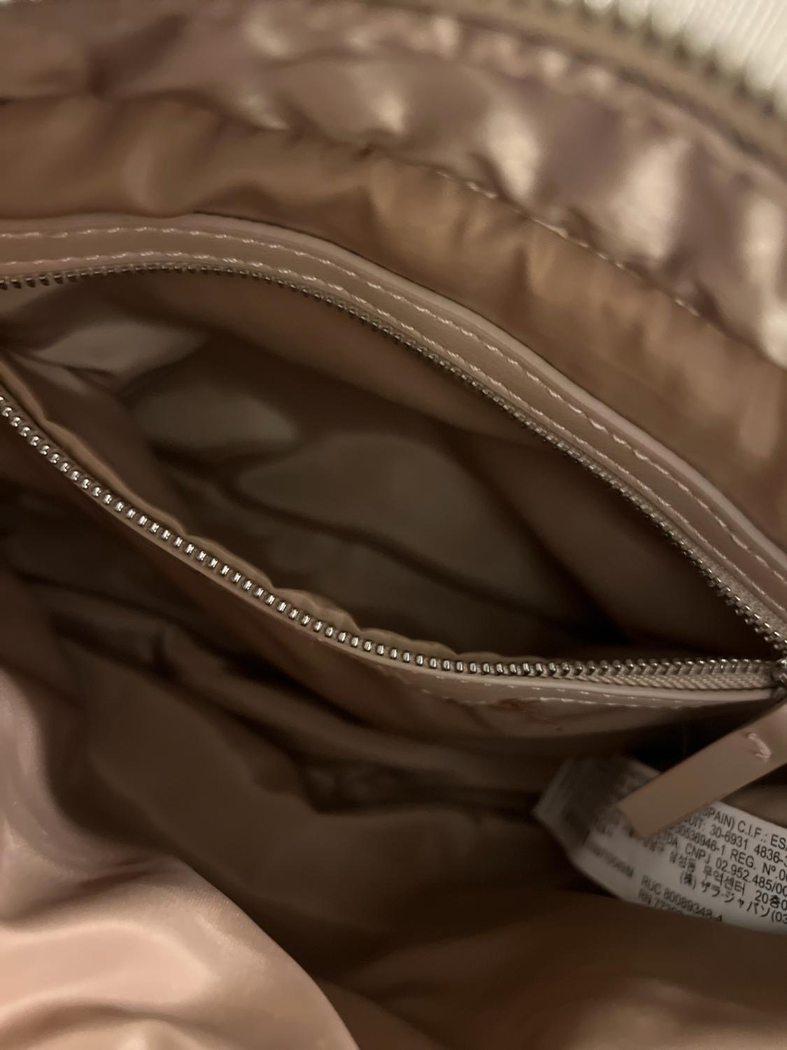 Detail of the inside of a beige handbag. An interior zippered pocket and a white label with text are visible.