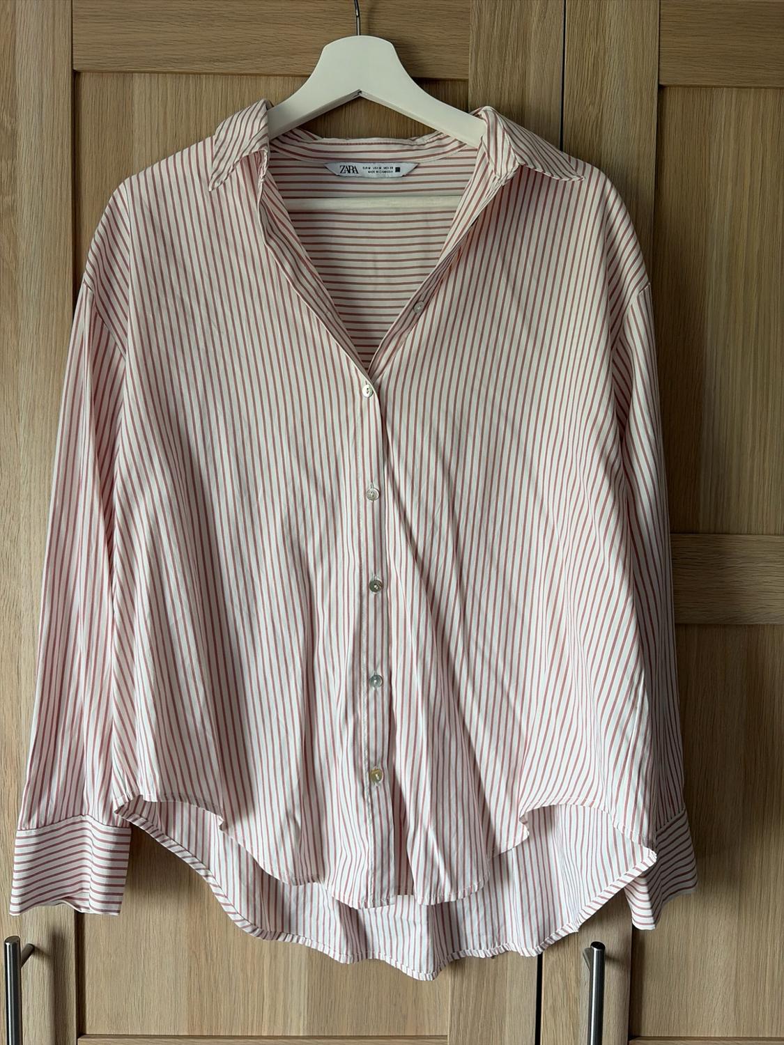 Pink and white striped long-sleeved shirt hanging on a hanger in front of a wooden wardrobe. Label with text ZARA.