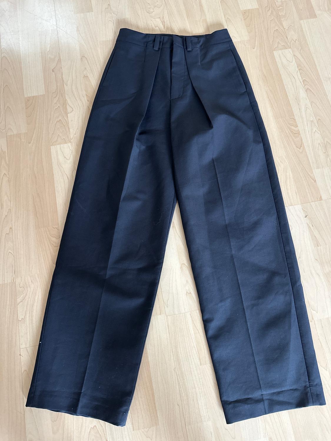Plain black fabric trousers with pleats, laid on a light wooden floor.
