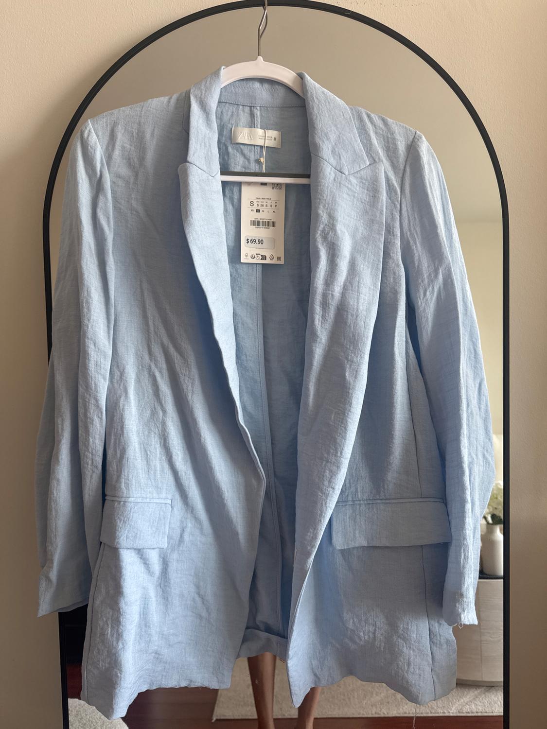 Light blue blazer hanging on a hanger in front of a mirror. Visible tag with price of $69.90. Neutral room background.