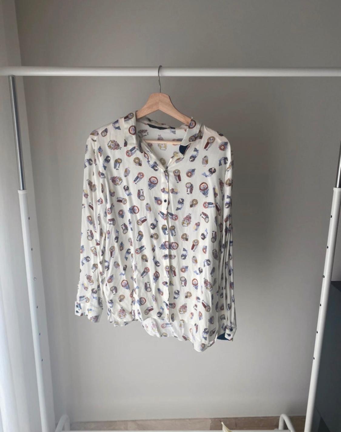 White shirt with a watch and owl print, hanging on a white clothes rack in a grey-walled room.