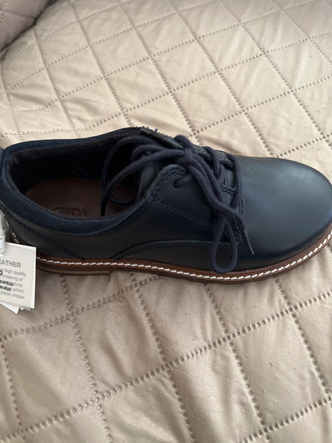 Black leather lace-up shoe on a quilted beige bedspread. Visible tag with partial English text.