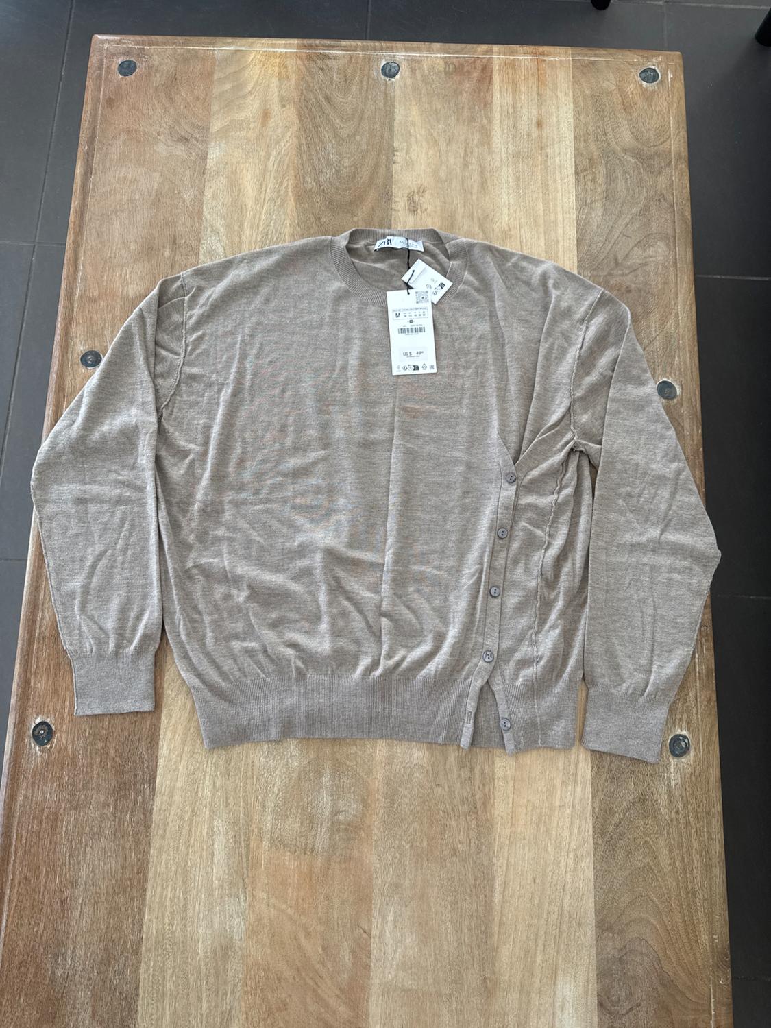 Long-sleeved beige jumper with side buttons on a wooden table. Hanging tags visible on the collar.