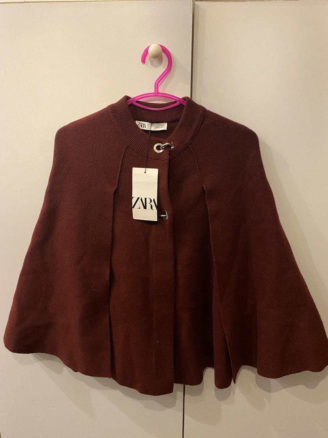 Brown cape hanging on a pink hanger with a visible Zara tag, against a light background.