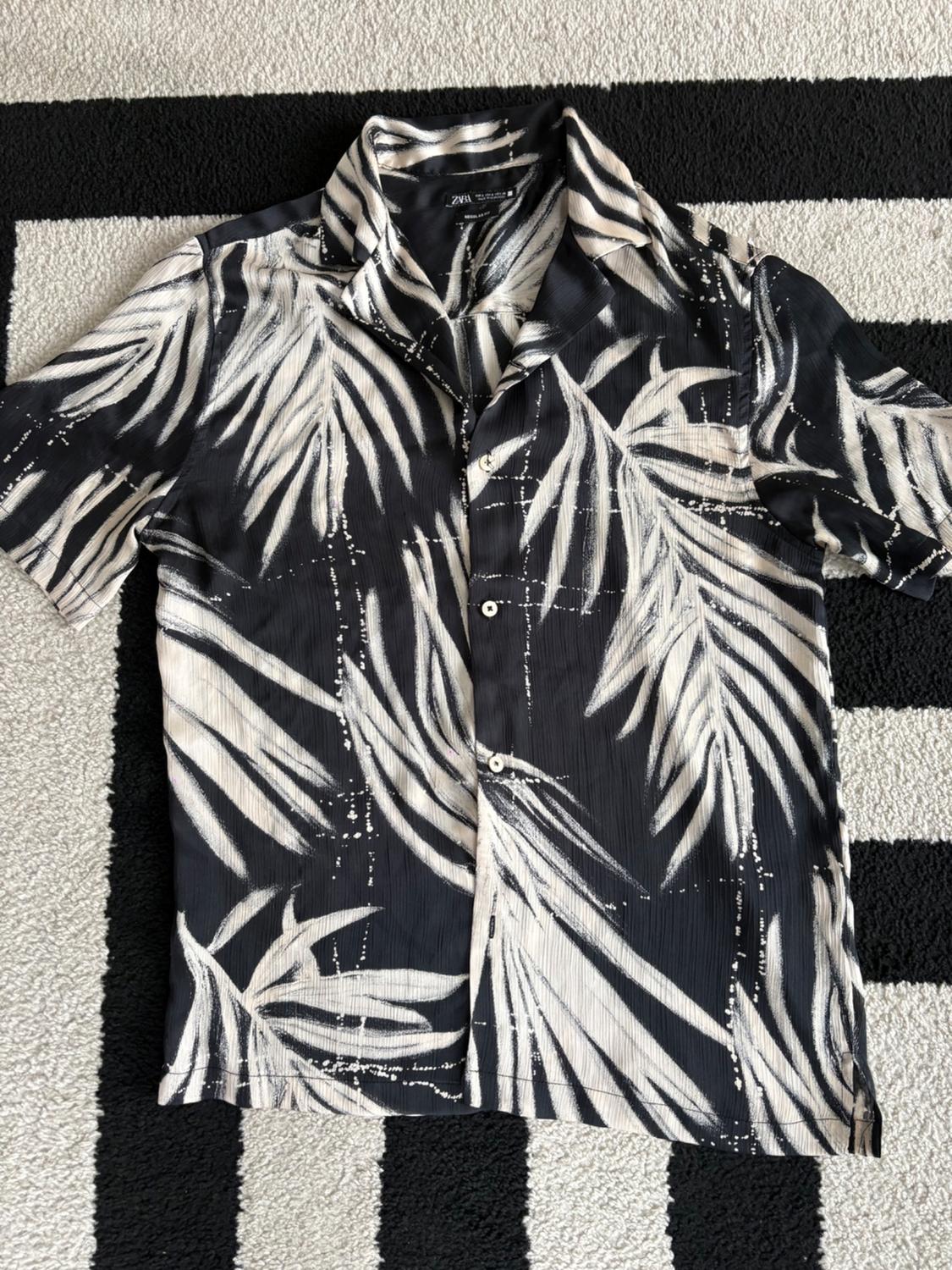 Short-sleeved shirt with a white leaf print on a black background, placed on a black and white striped rug.