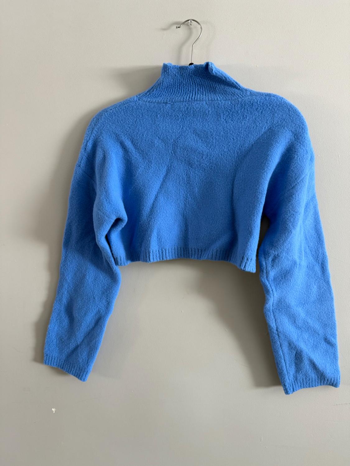 Blue turtleneck sweater hanging on a hanger against a beige wall. Long sleeves and a cropped design.