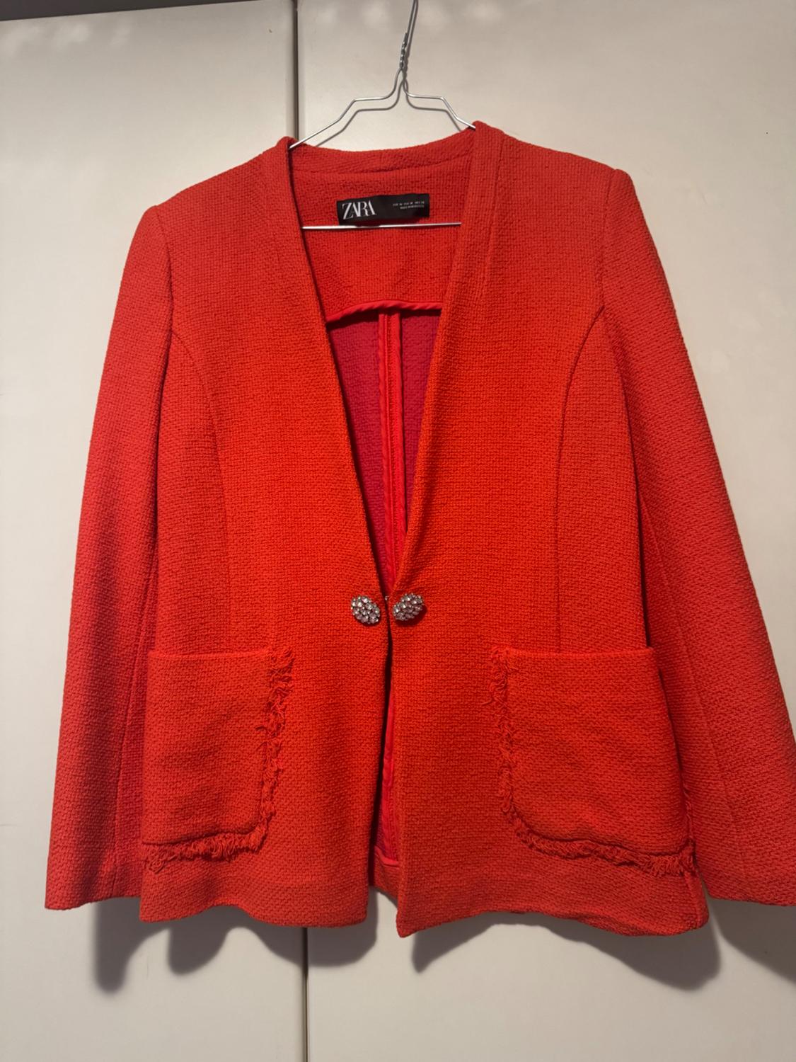 Red textured jacket, two front pockets and shiny button fastening, hanging on a hanger. Visible label: ZARA.