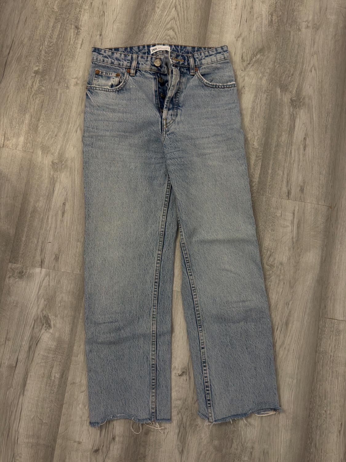 Ανοιχτό μπλε ίσιο τζιν παντελόνι με ετικέτα ZARA, κούμπωμα με κουμπιά και ξεφτισμένο τελείωμα, σε γκρι ξύλινο πάτωμα.