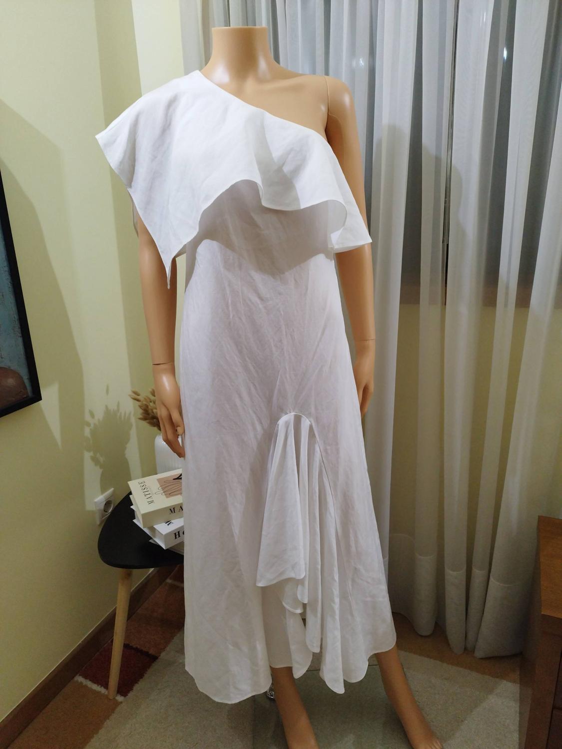 Mannequin wearing a white one-shoulder dress with frills. Interior with curtains and a table with books bearing the names MATISSE, MA, H.