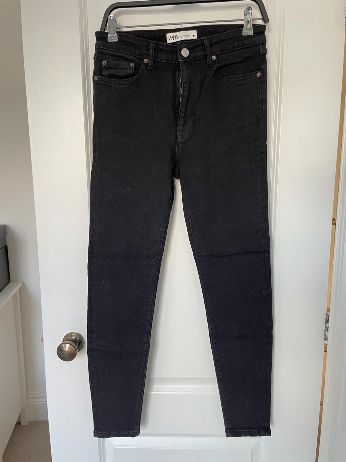 Black trousers hanging on a white door, with a visible label on the waistband. Indoor setting with natural light.