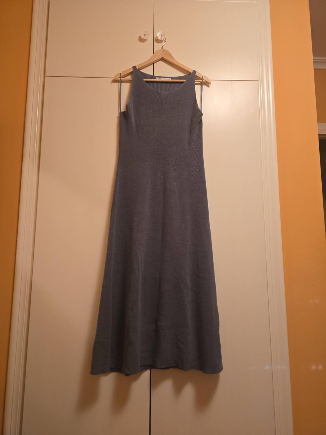 Sleeveless long blue-grey knitted dress hanging on a wooden hanger above a white wardrobe in a room.