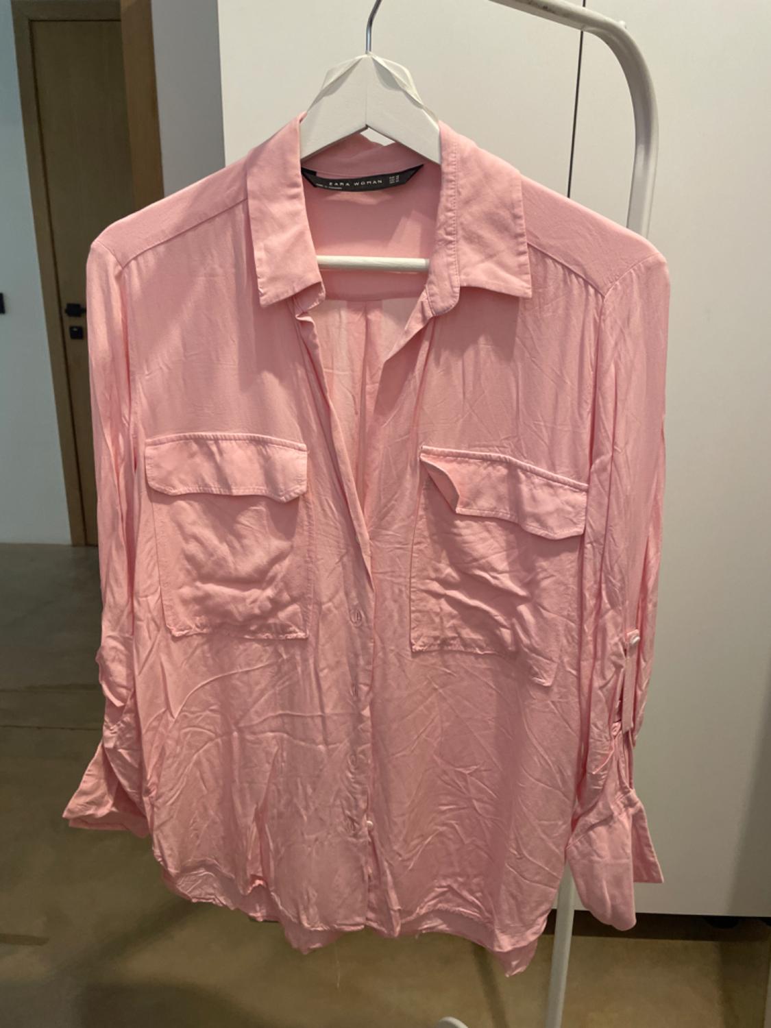 Pink long-sleeved shirt with front pockets, hanging on a white hanger. Interior room background.