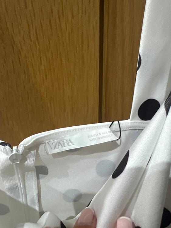 Label of a white garment with black polka dots, showing the brand "ZARA" and "MADE IN MOROCCO" on a light wood background.
