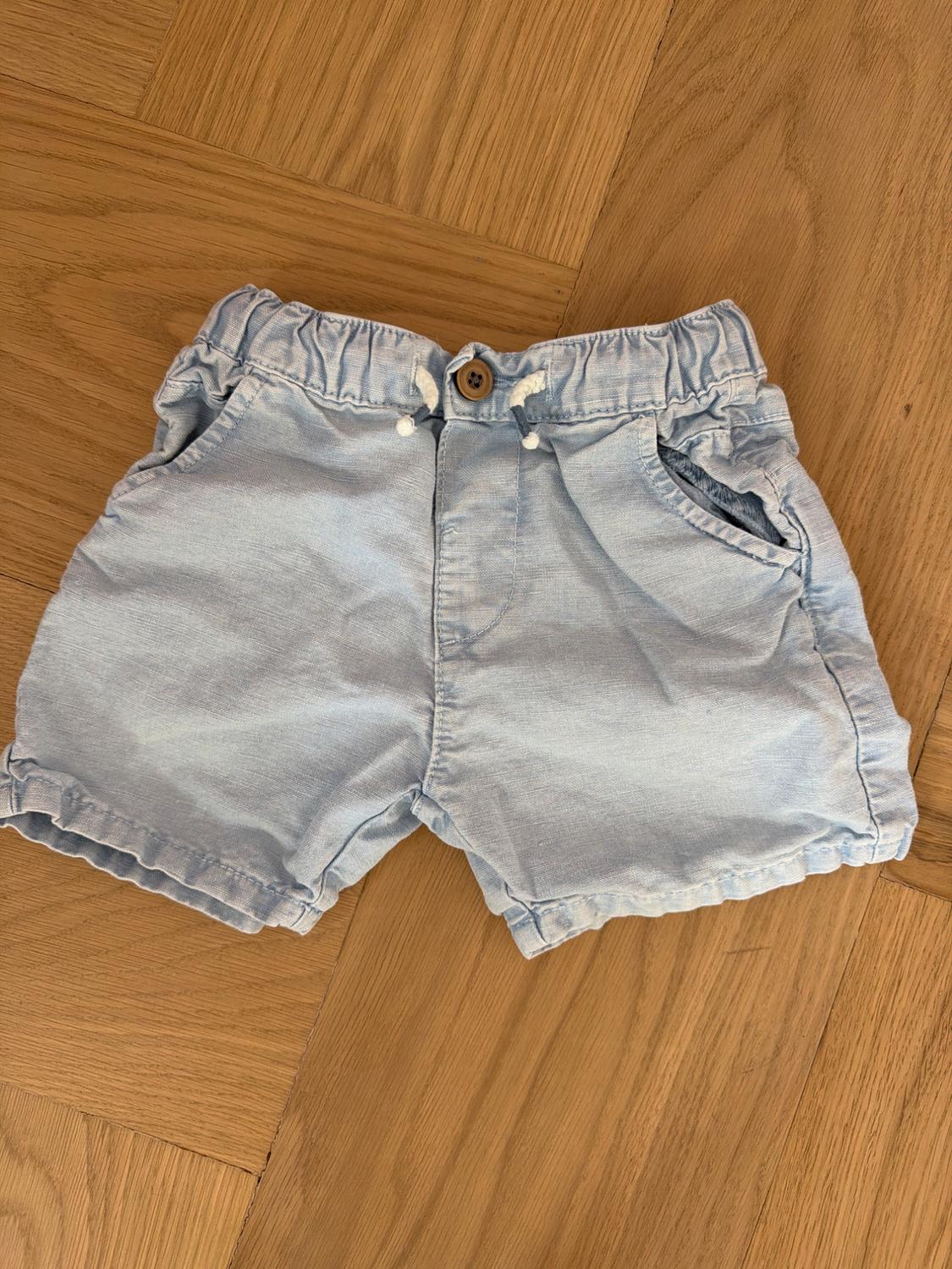 Light blue, lightweight fabric shorts with an elasticated waist and a wooden button, on a light wooden floor.
