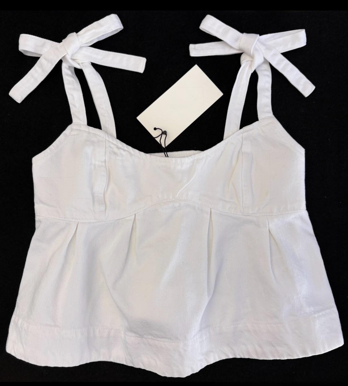 White strappy top with bows and stitching on the chest, on a black background. It has a white tag hanging.