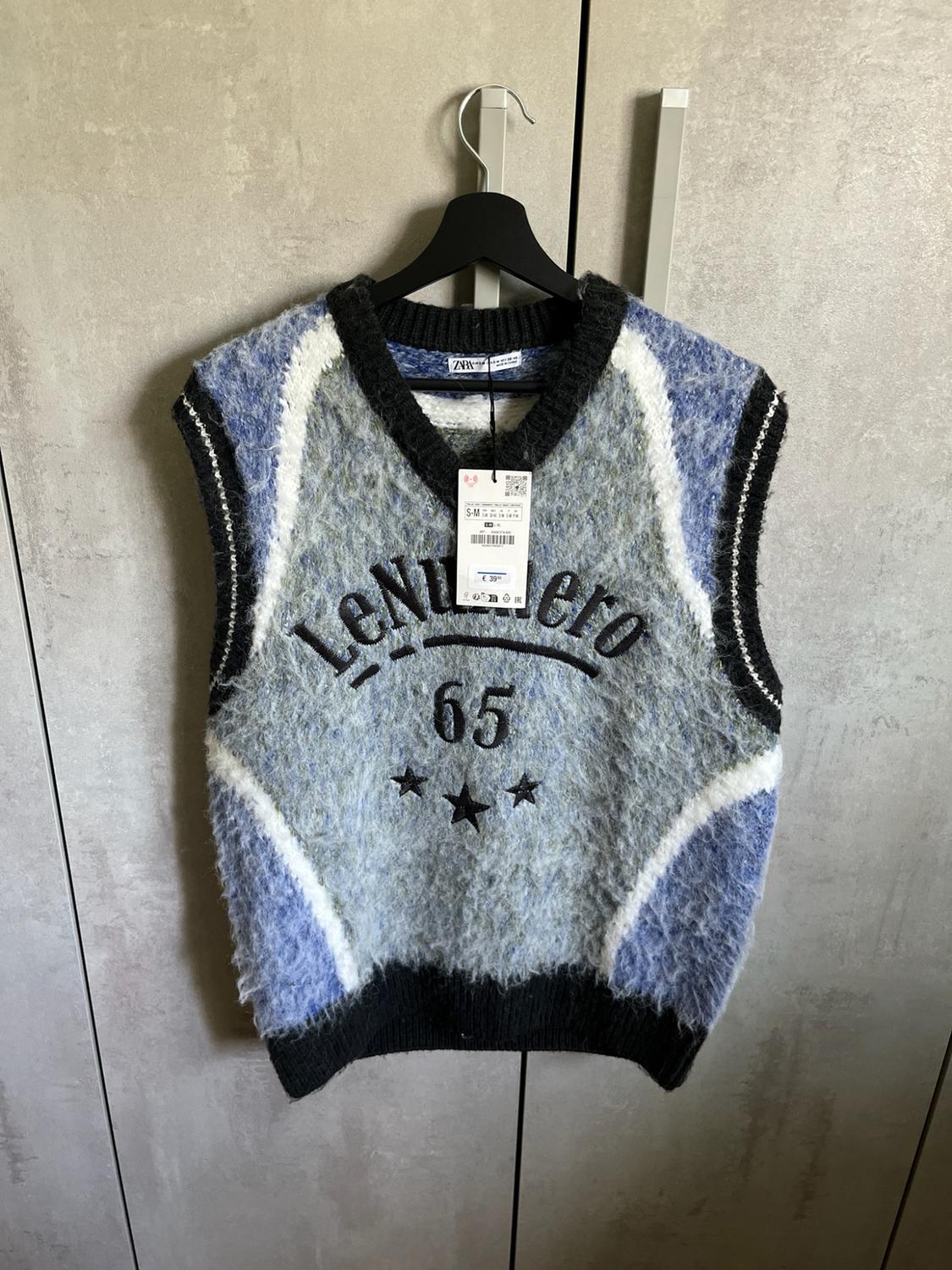 Knitted waistcoat in shades of grey, blue and white hanging in a wardrobe. Includes the text LeNumero 65 and three black stars.