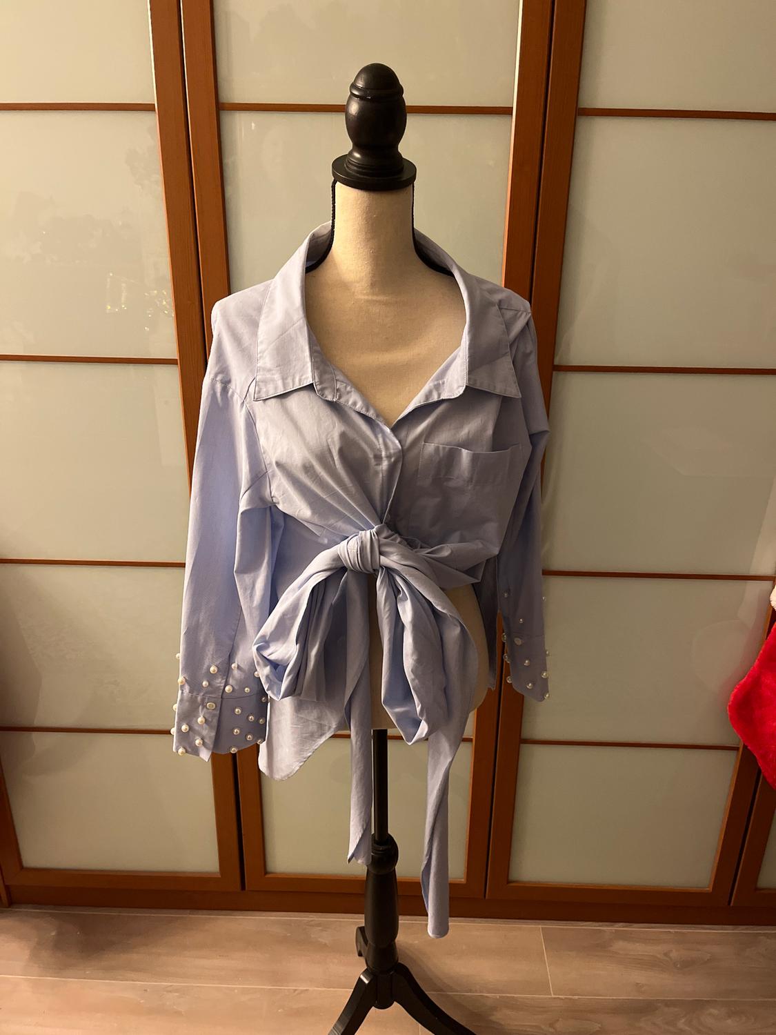 Mannequin with a light blue blouse knotted at the front, pearl details on the sleeves, wooden and glass sliding doors in the background.