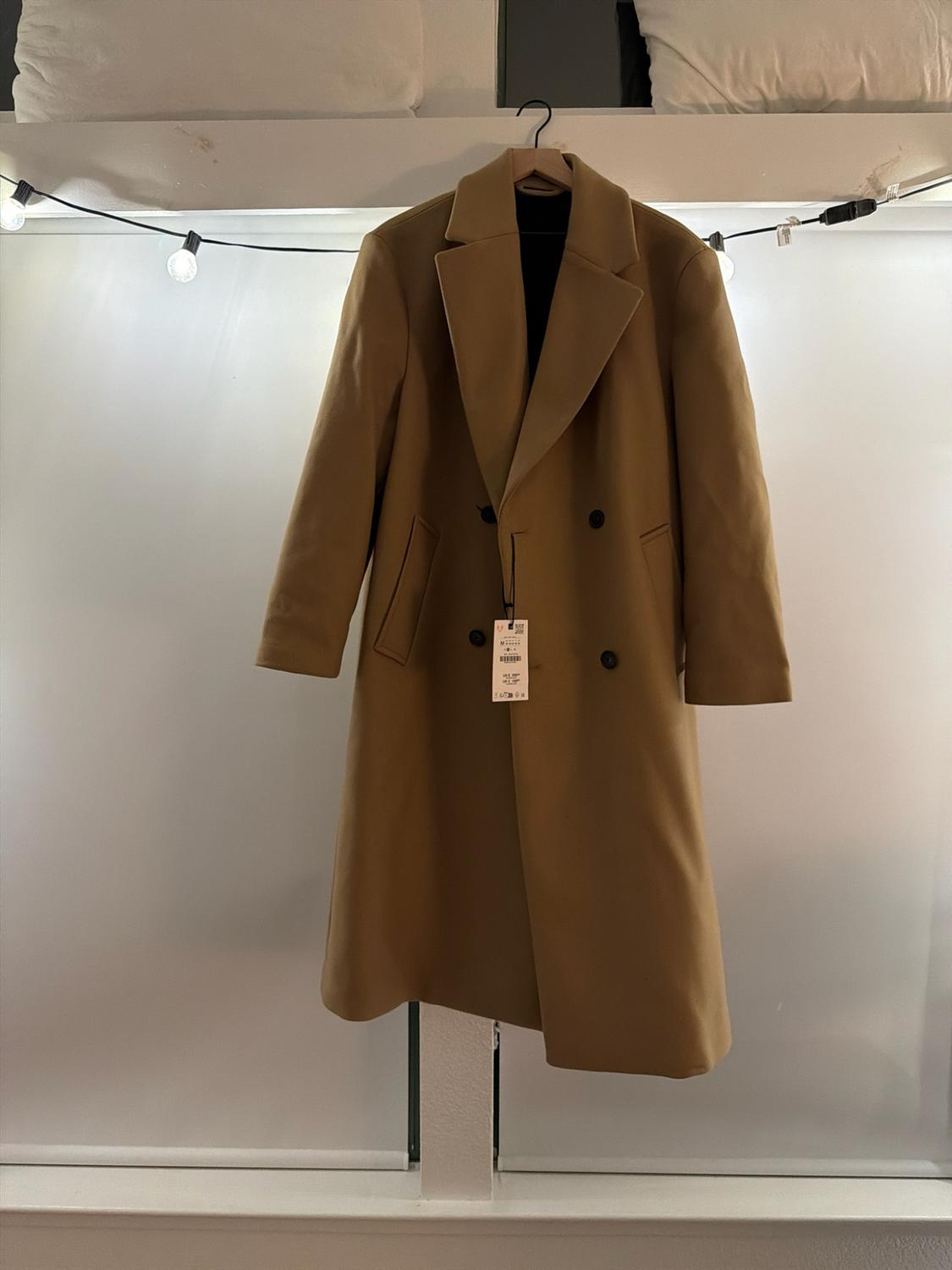 Long camel-colored double-breasted coat with black buttons, hanging on a hanger in front of a white wall with a string of lights.