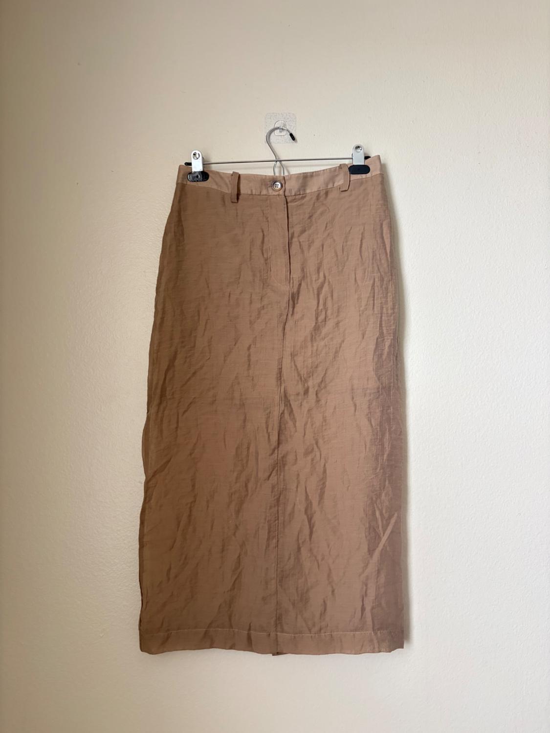 Light brown straight maxi skirt with a crinkled texture, belt loops and a front button. Hanging on a transparent hanger against a light-coloured wall.