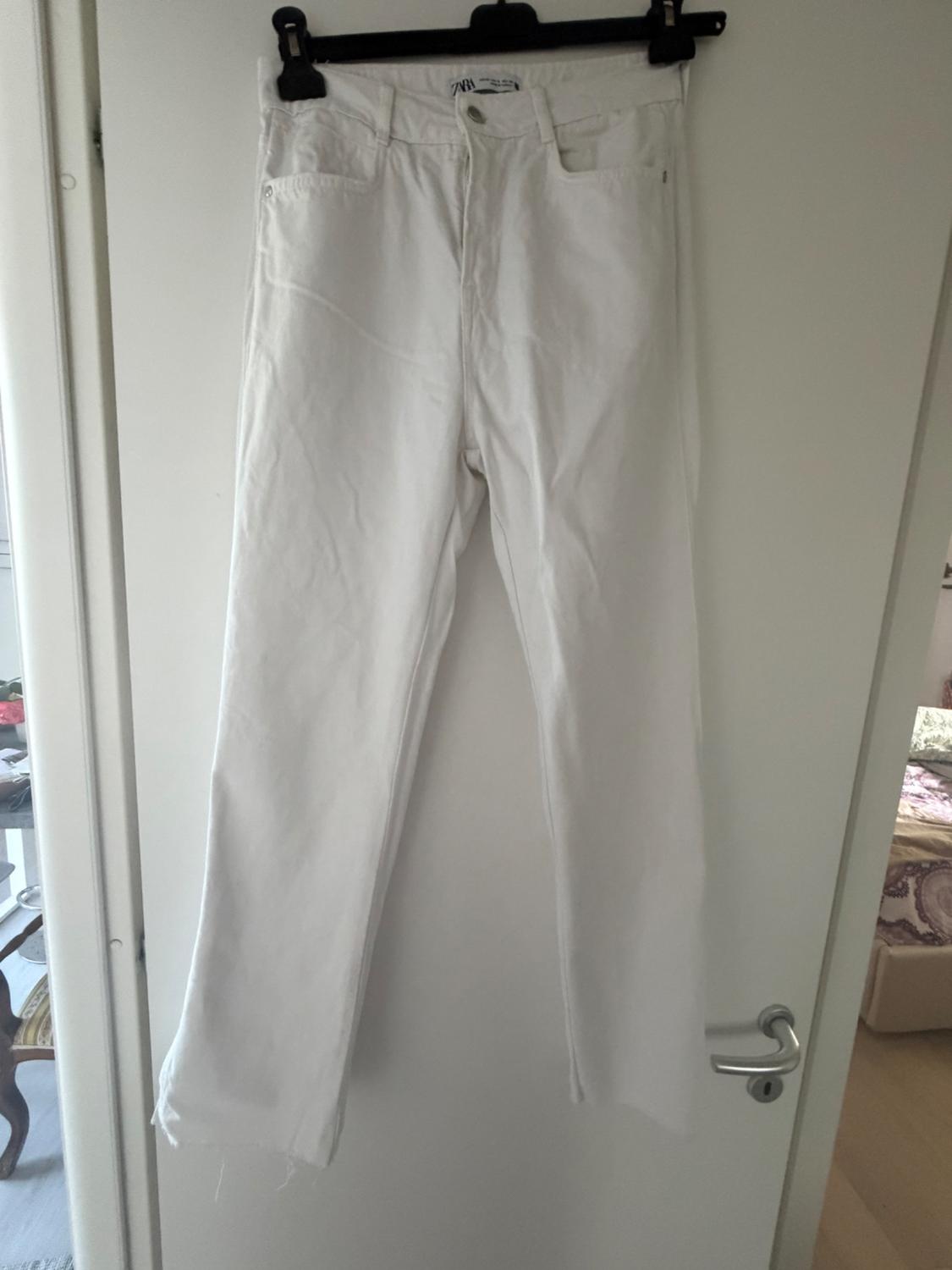 White trousers with a frayed hem hanging on a black hanger over a white door in a domestic setting. Label with the text ZARA.