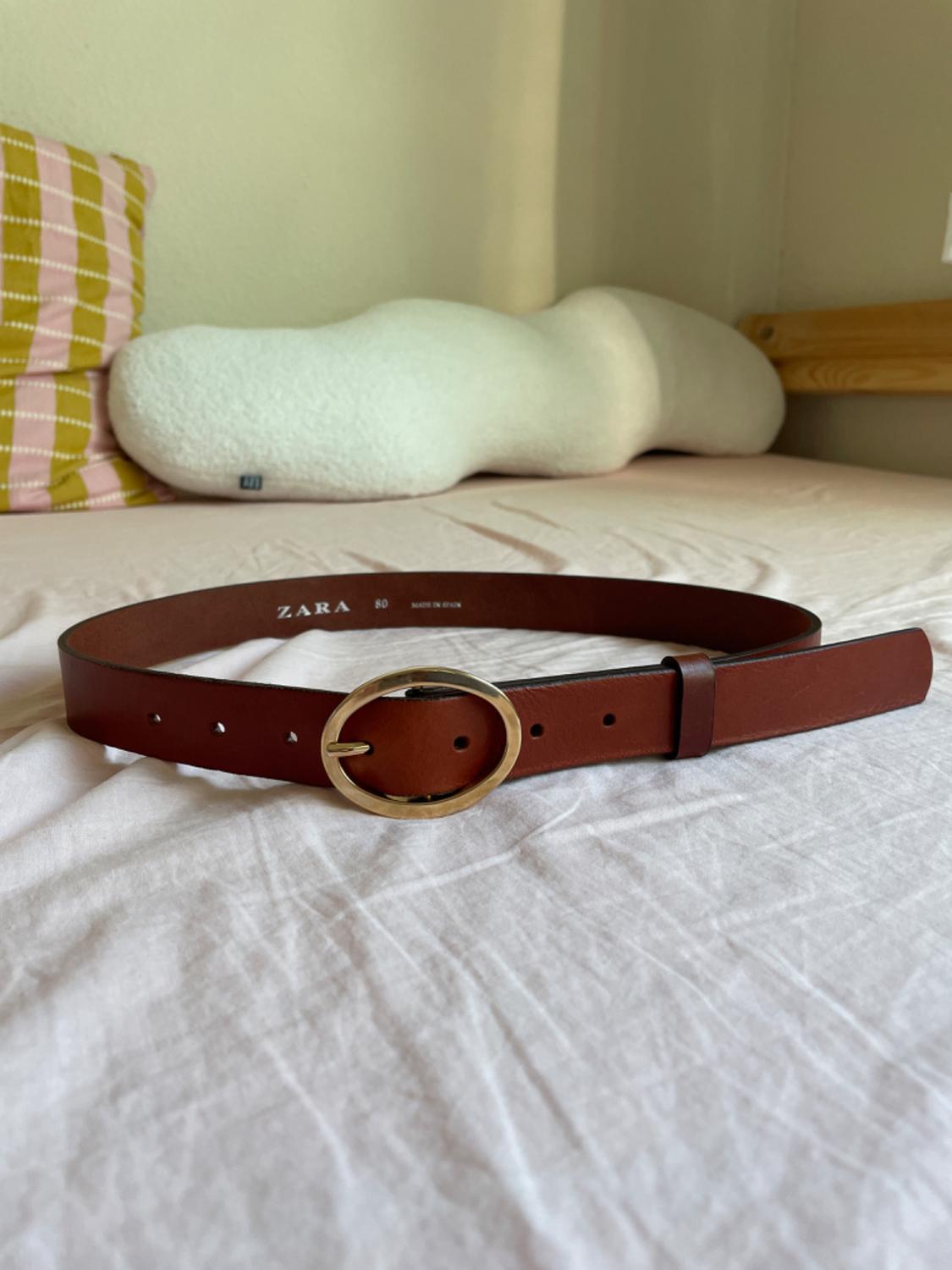 Brown belt with a gold oval buckle on a bed with white sheets. White pillow and striped cushion in the background. Text: ZARA 80.
