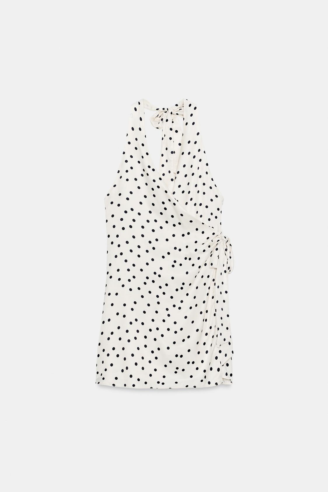 Front view of a short, ecru dress with black polka dots, a halter neck, a V-neckline, and a crossover side fastening with a tie.