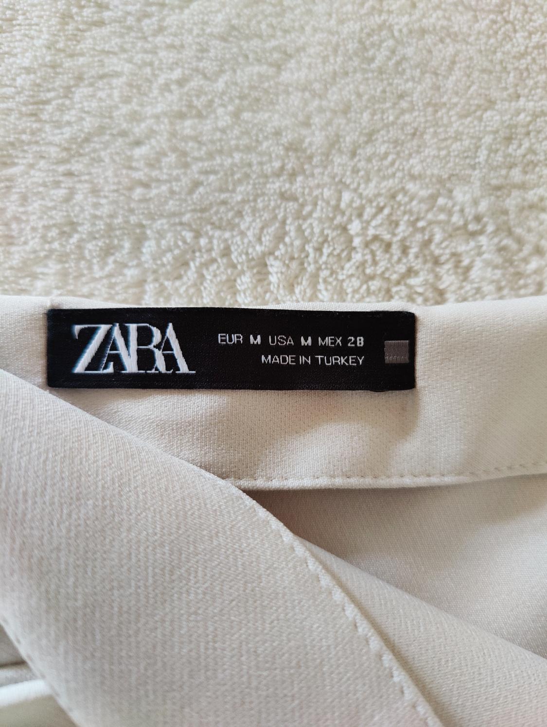 Μαύρη ετικέτα Zara σε ένα μπεζ ρούχο, με το κείμενο "EUR M USA M MEX 28 MADE IN TURKEY", σε φόντο από λευκό υφασμάτινο φόντο με υφή.