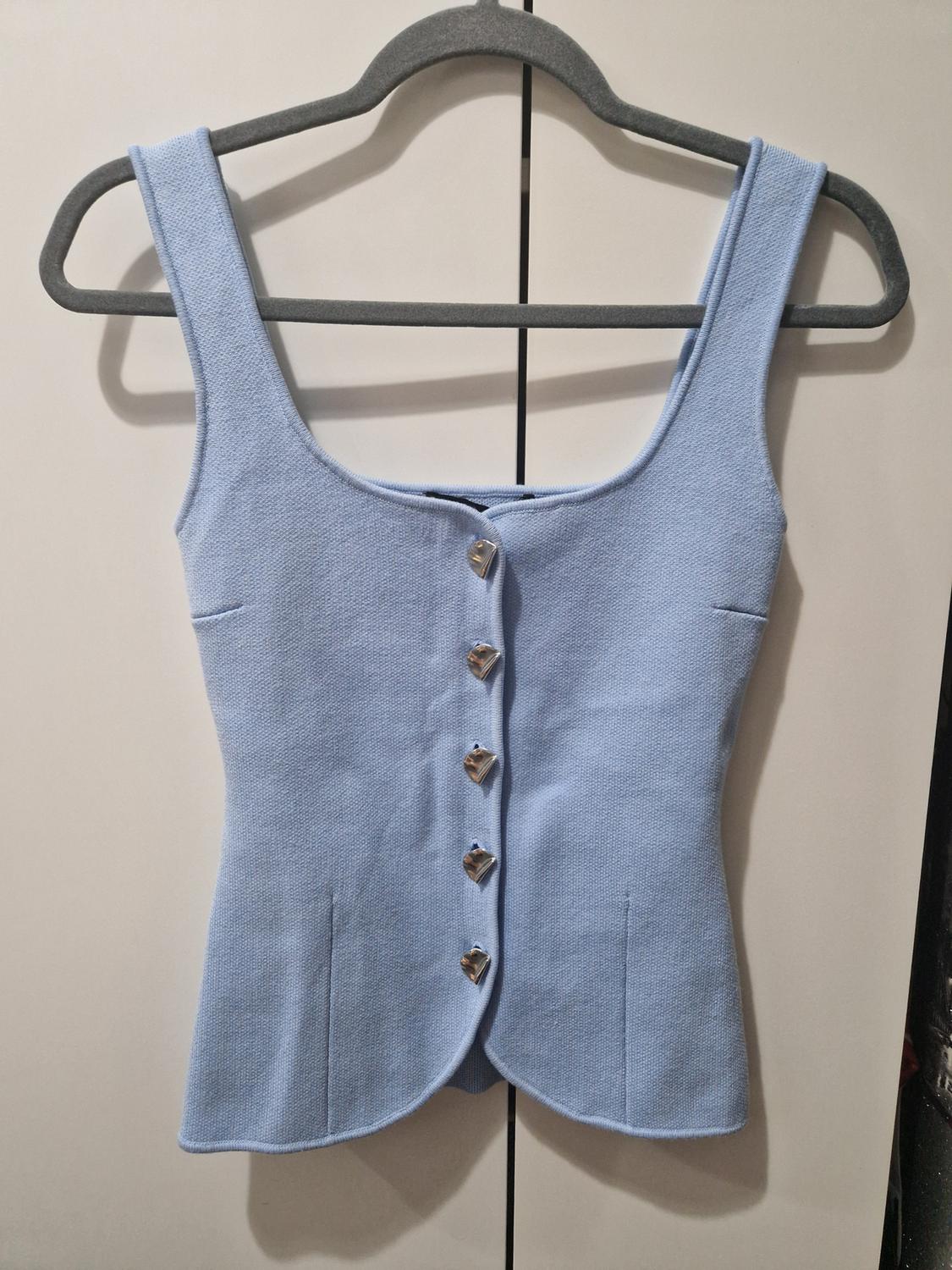 Light blue textured top with a square neckline and silver buttons, hanging on a grey hanger in front of a white wardrobe.