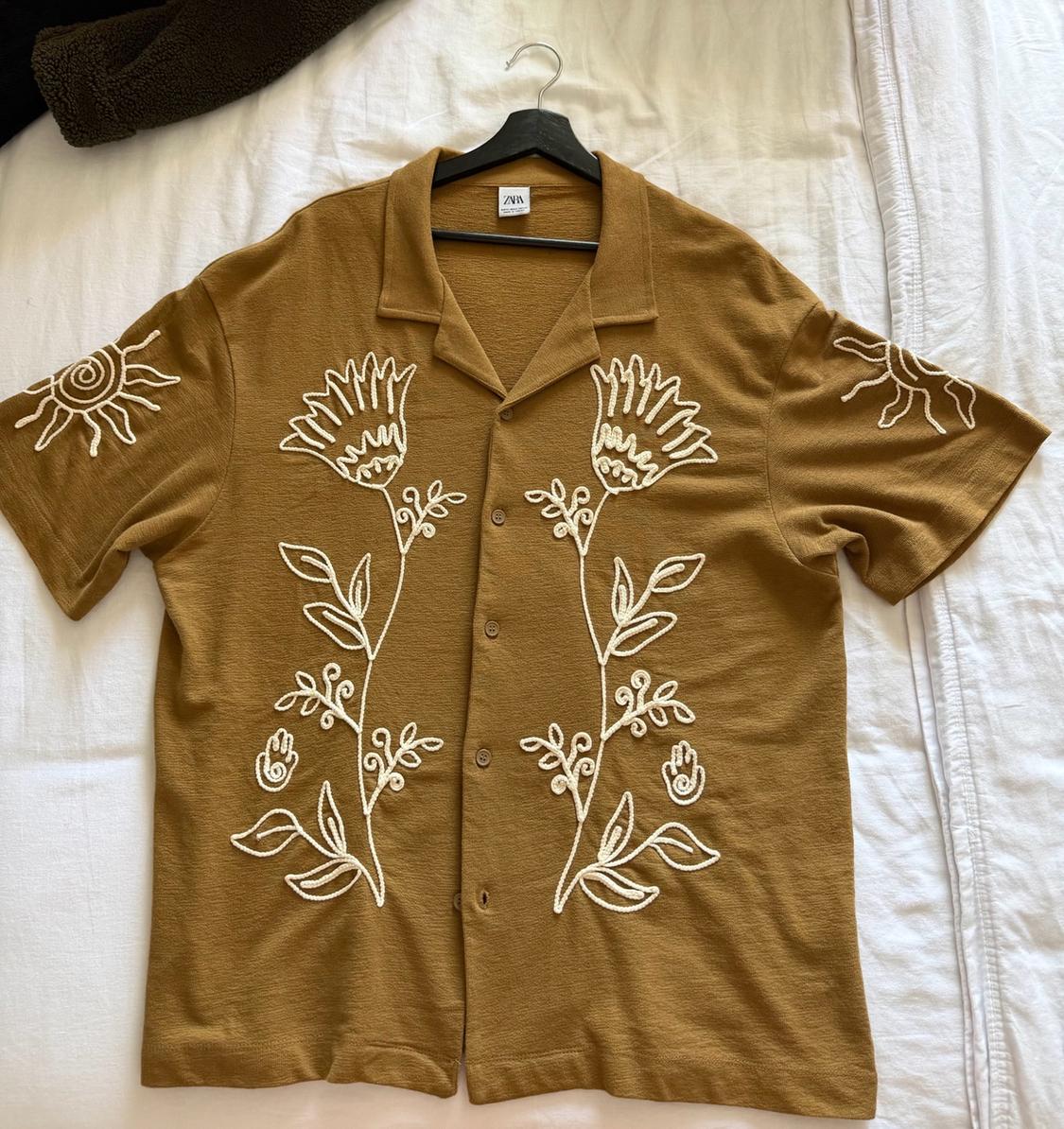 Mustard short-sleeved shirt with white floral and sun embroidery on white bed sheets. Black hanger and tag with visible ZARA text.
