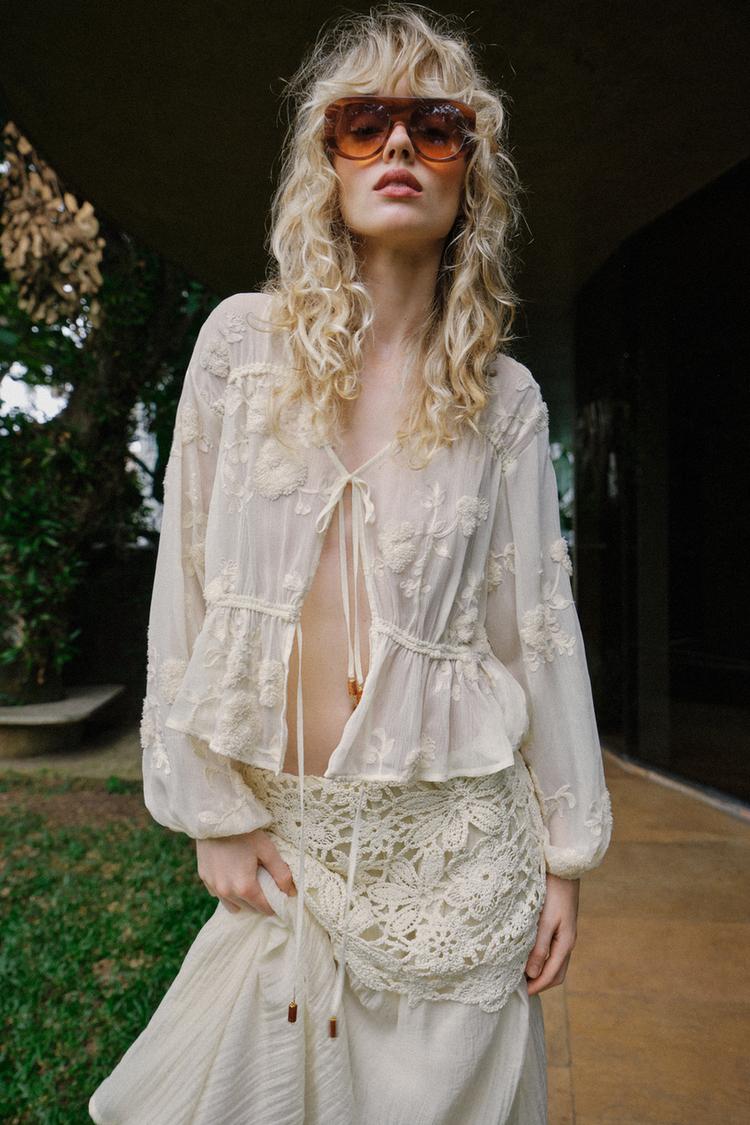 Poloviční záběr na světle béžovou průsvitnou halenku s květinovou výšivkou, výstřihem do V, dlouhými rukávy a zavazováním s korálky.