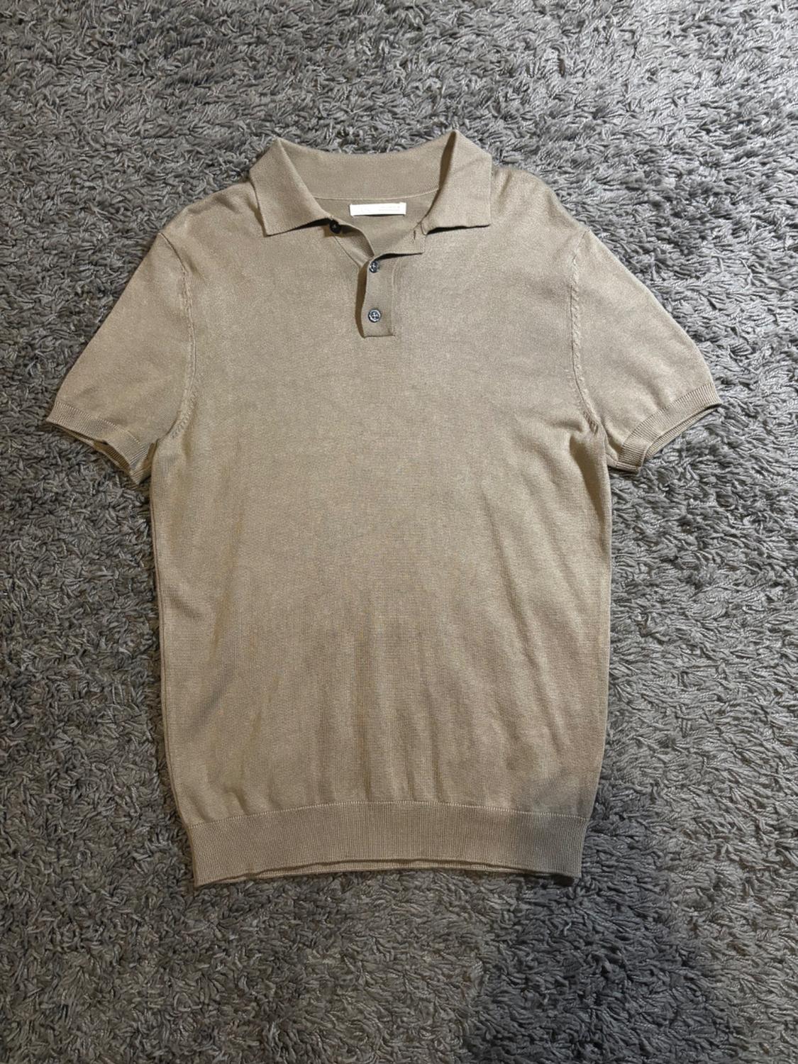 Beige short-sleeved polo shirt with two buttons, placed on a thick-textured grey rug.