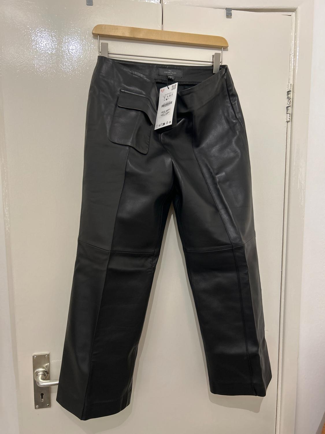Black leather trousers hanging on a wooden hanger over a white door, with a visible label on the pocket.