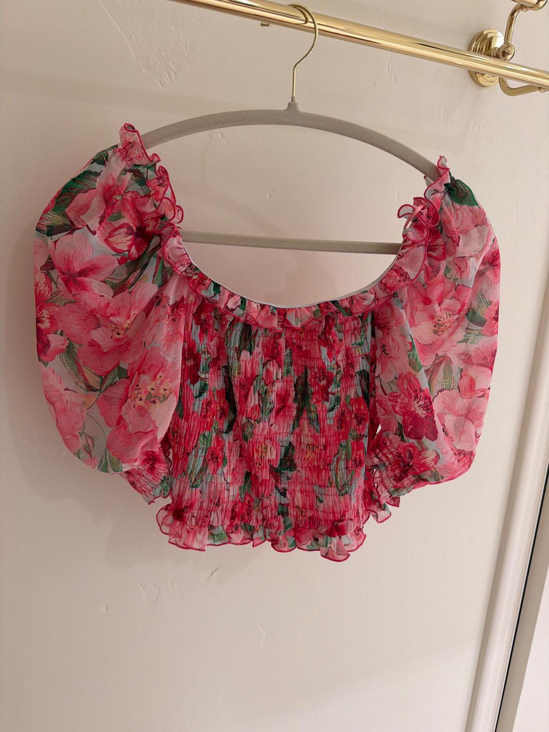Short chiffon blouse with a floral print in pink and green tones, hanging on a gold hanger against a white wall.