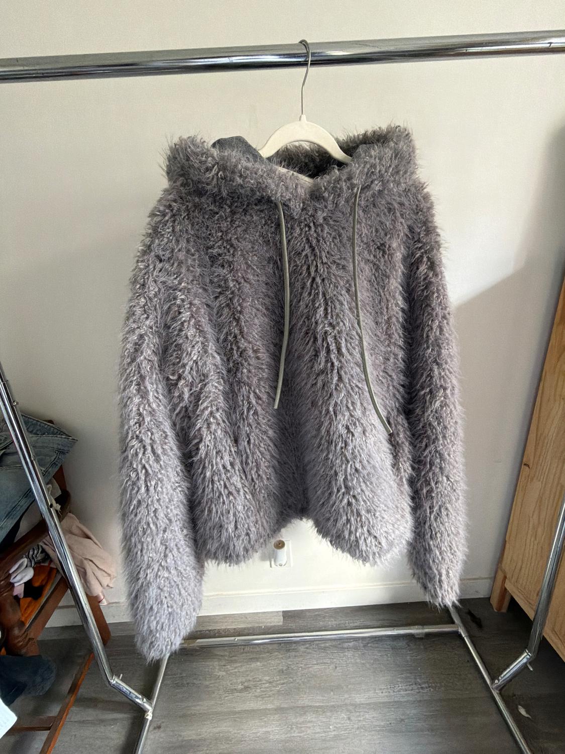 Grey fleece hoodie with drawstrings, hanging on a metal clothes rail, on a light wooden floor.