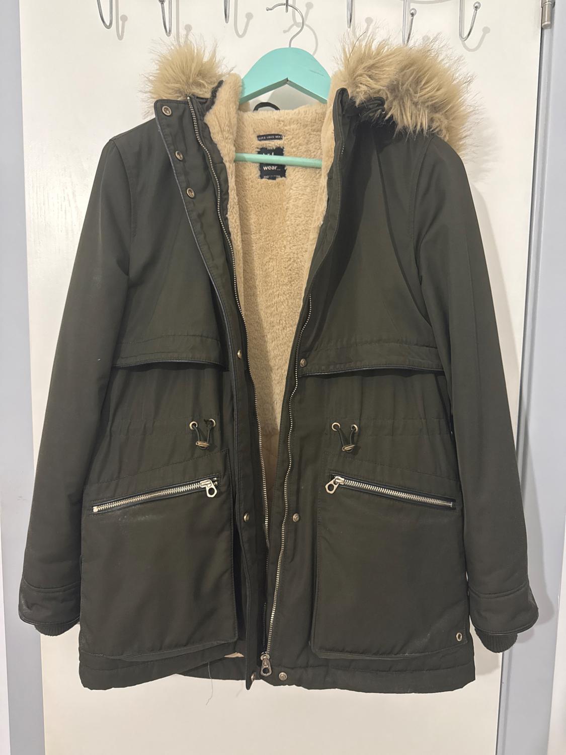 Olive green coat with a faux fur hood and beige lining, hanging on a blue hanger on a white door.