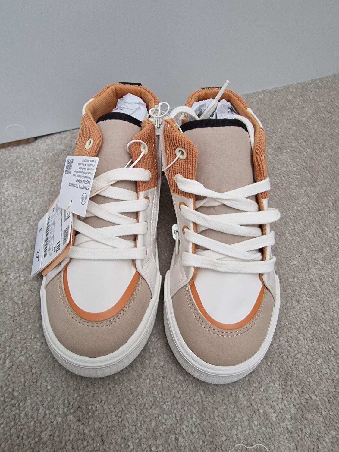 Beige and orange high-top trainers with white laces on a grey carpet. Tags hanging from one shoe.