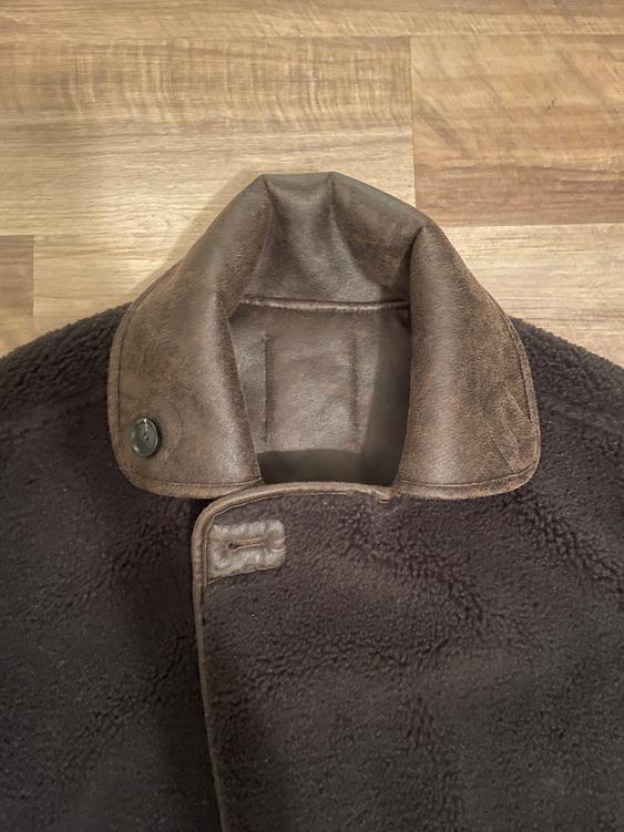 Dark brown wool coat with a brown leather collar on a light wooden background. Button detail on the collar.