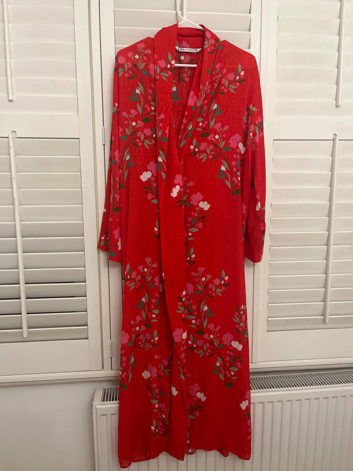 Long red dress with a floral print in shades of green, pink and white, hanging on a hanger in front of white blinds.