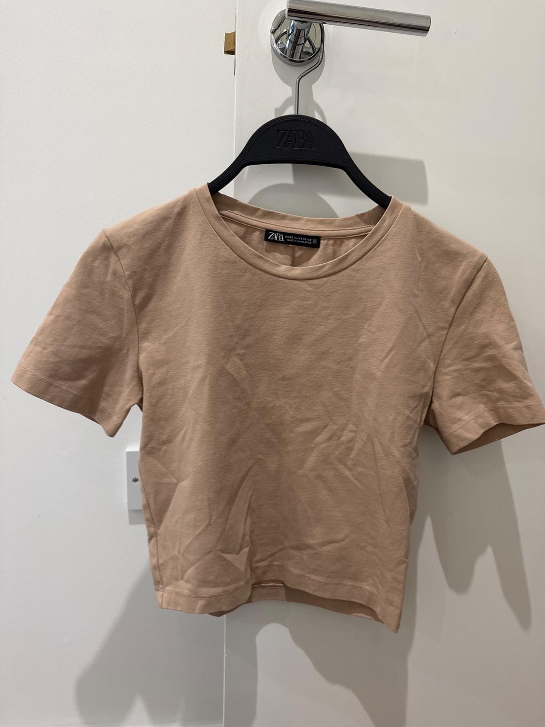 A beige short-sleeved T-shirt hanging on a black hanger with the brand visible, over a white door.