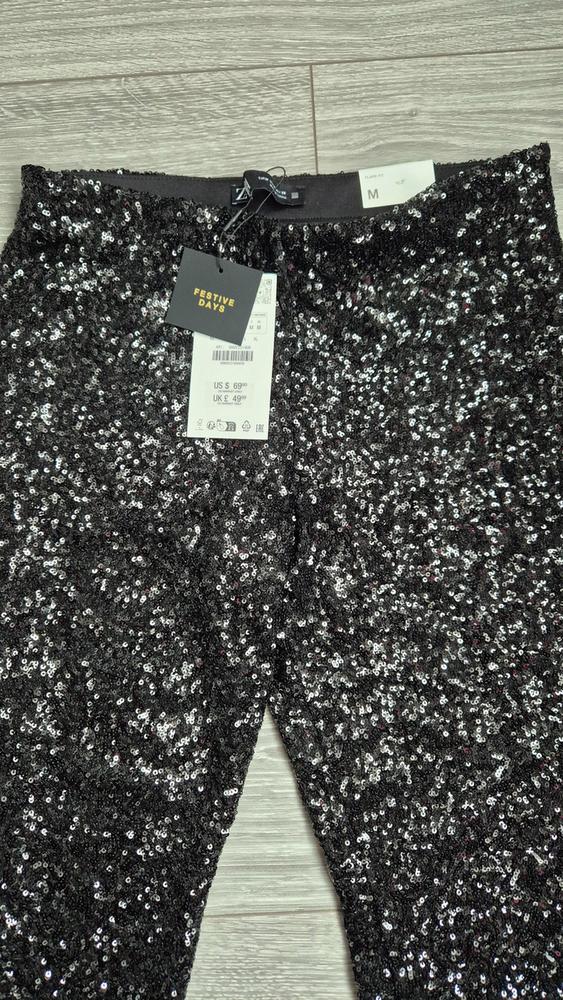 Black sequinned trousers on a light wooden floor. Visible label with text "FESTIVE DAYS" and sizes M, US S, UK £49.99.
