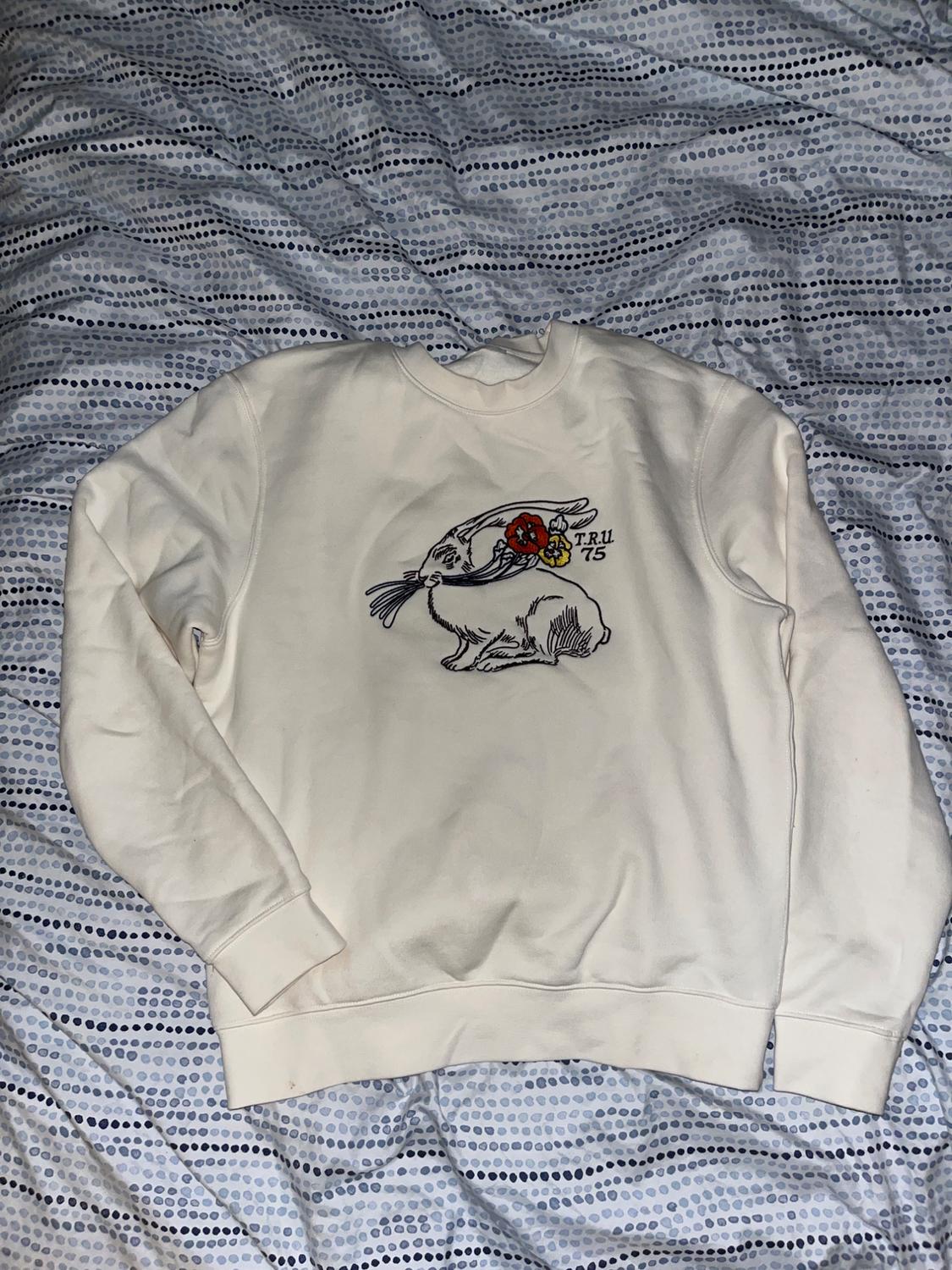 White sweatshirt with a bird and floral design on the chest, text "TRU 75", on a bed with a blue and white polka dot bedspread.