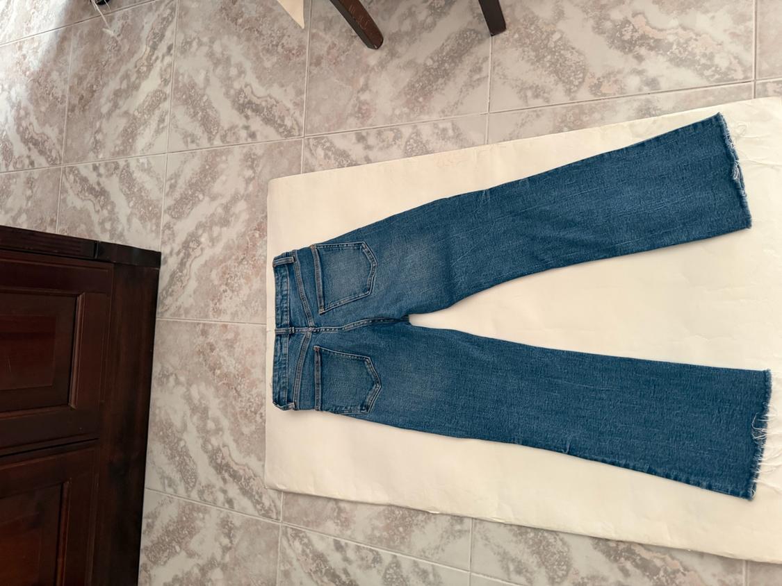 Blue jeans laid out, viewed from behind with a frayed hem, on a white background on a tiled floor next to a dark piece of furniture.