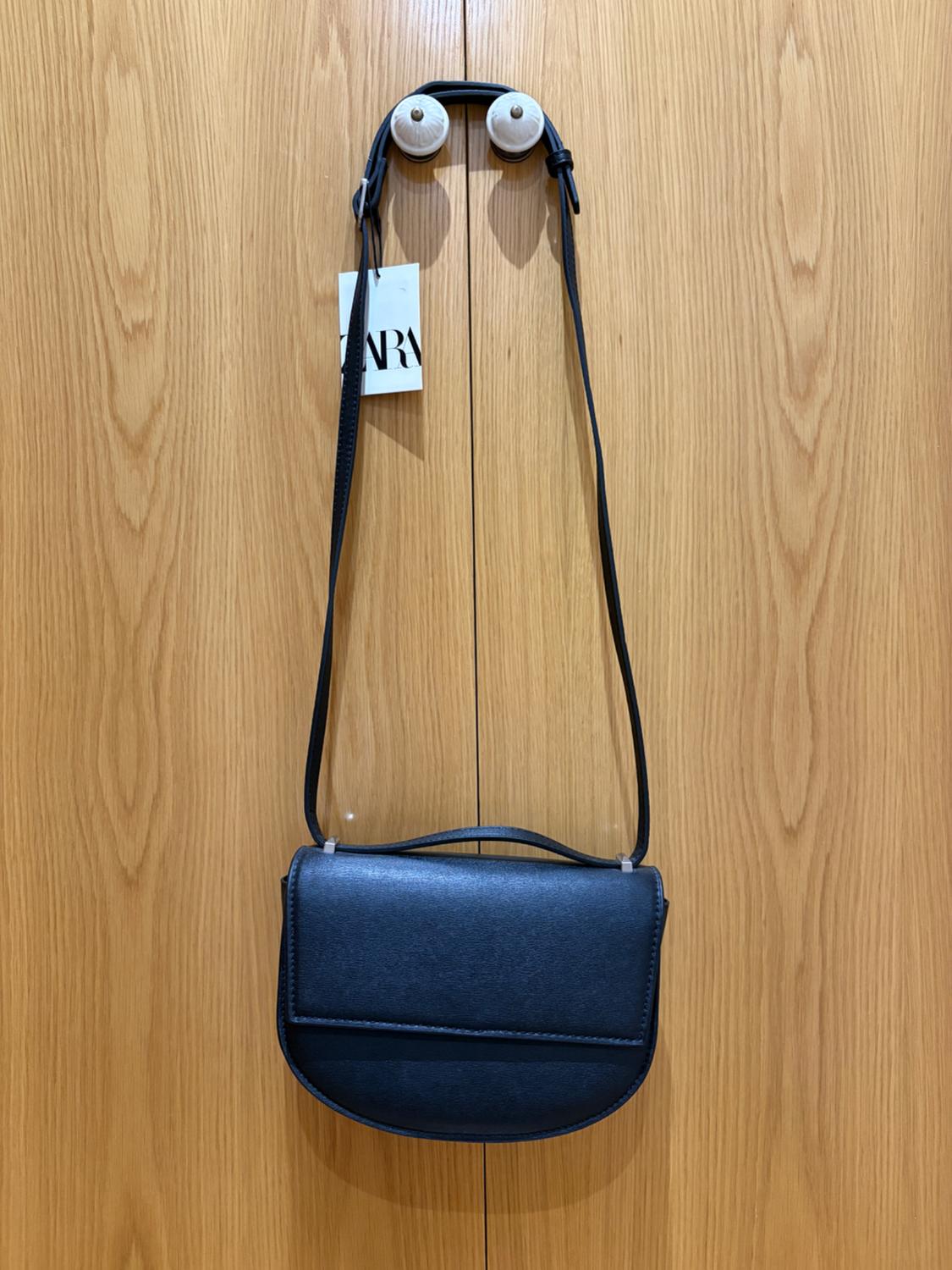Black handbag hanging on a light wooden door. Visible label with the text "ZARA". Simple and elegant design.