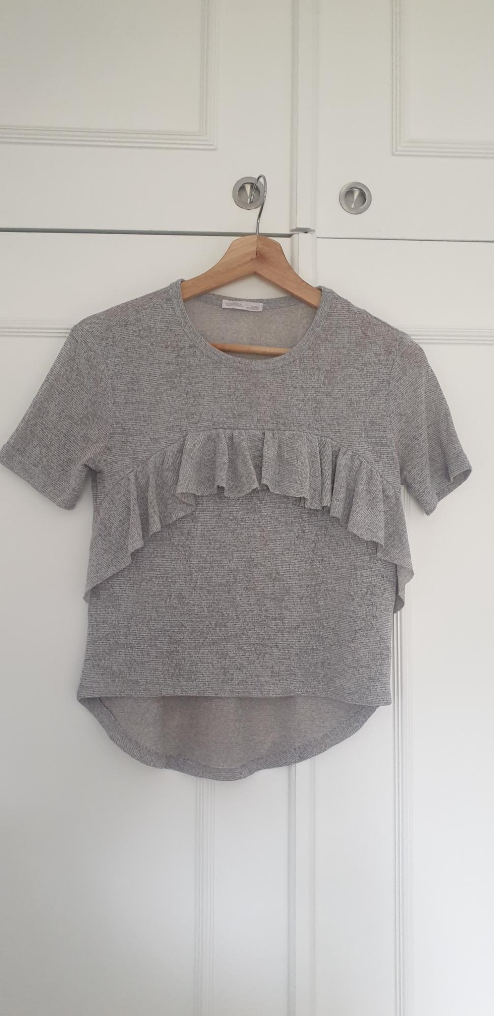 Grey T-shirt with ruffles on the chest, hanging on a wooden hanger in front of a white door.