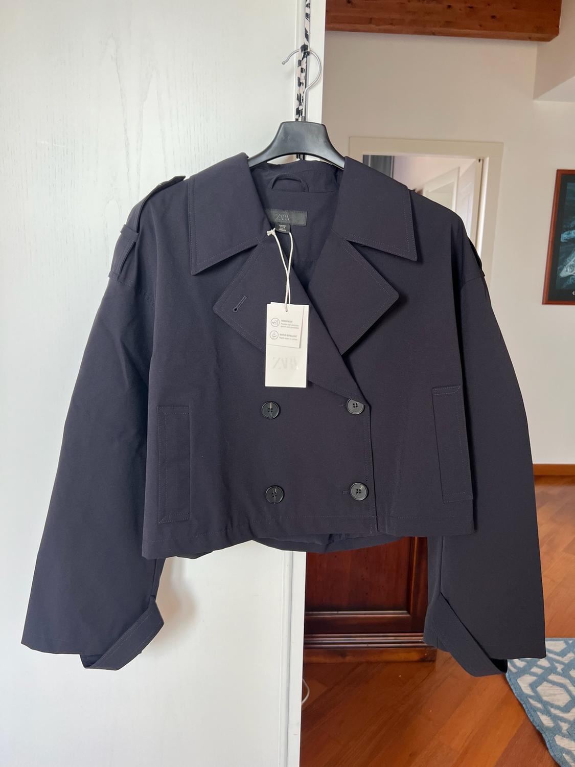 Short dark double-breasted jacket with wide lapels and buttons, hanging on a coat hanger in a room. White label with the text ZARA.