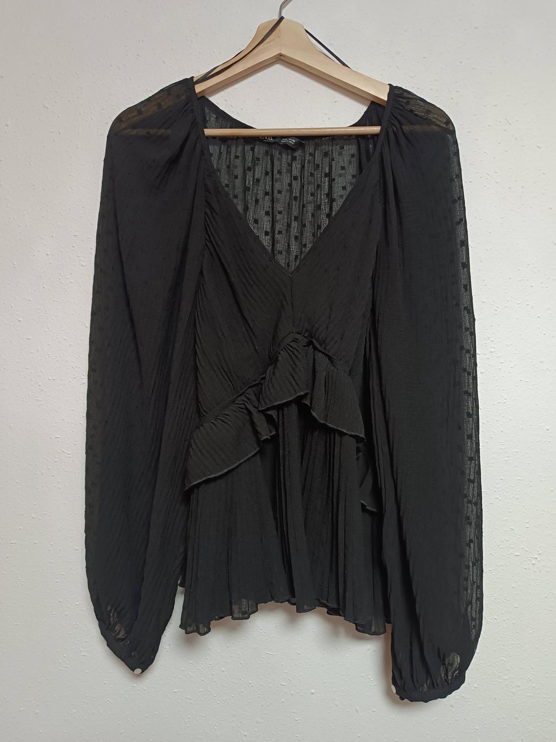 Black long-sleeved blouse with a pleated texture and ruffles on the front, hanging on a wooden hanger against a white background.