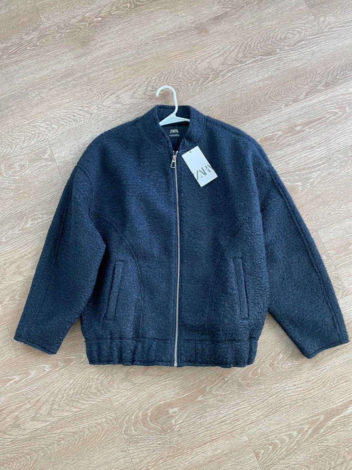Dark blue jacket with zipper and Zara tag, hanging on a white hanger over a light wood floor.