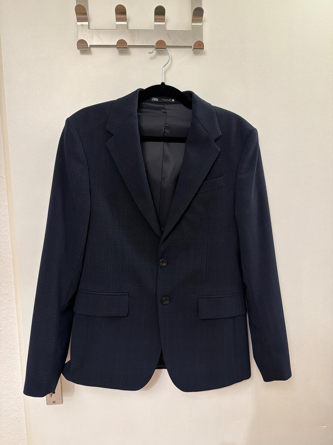 Dark blue jacket hanging on a black hanger on a metal wall hook. Plain white background.