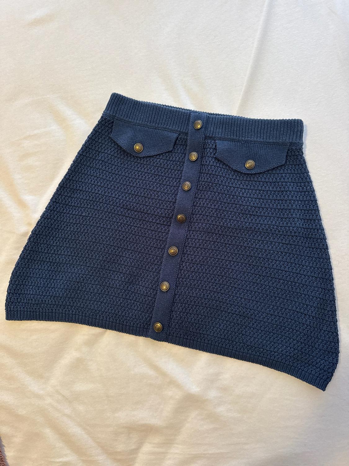 Navy blue knitted miniskirt with front buttons and two flap pockets, set against a white fabric background.