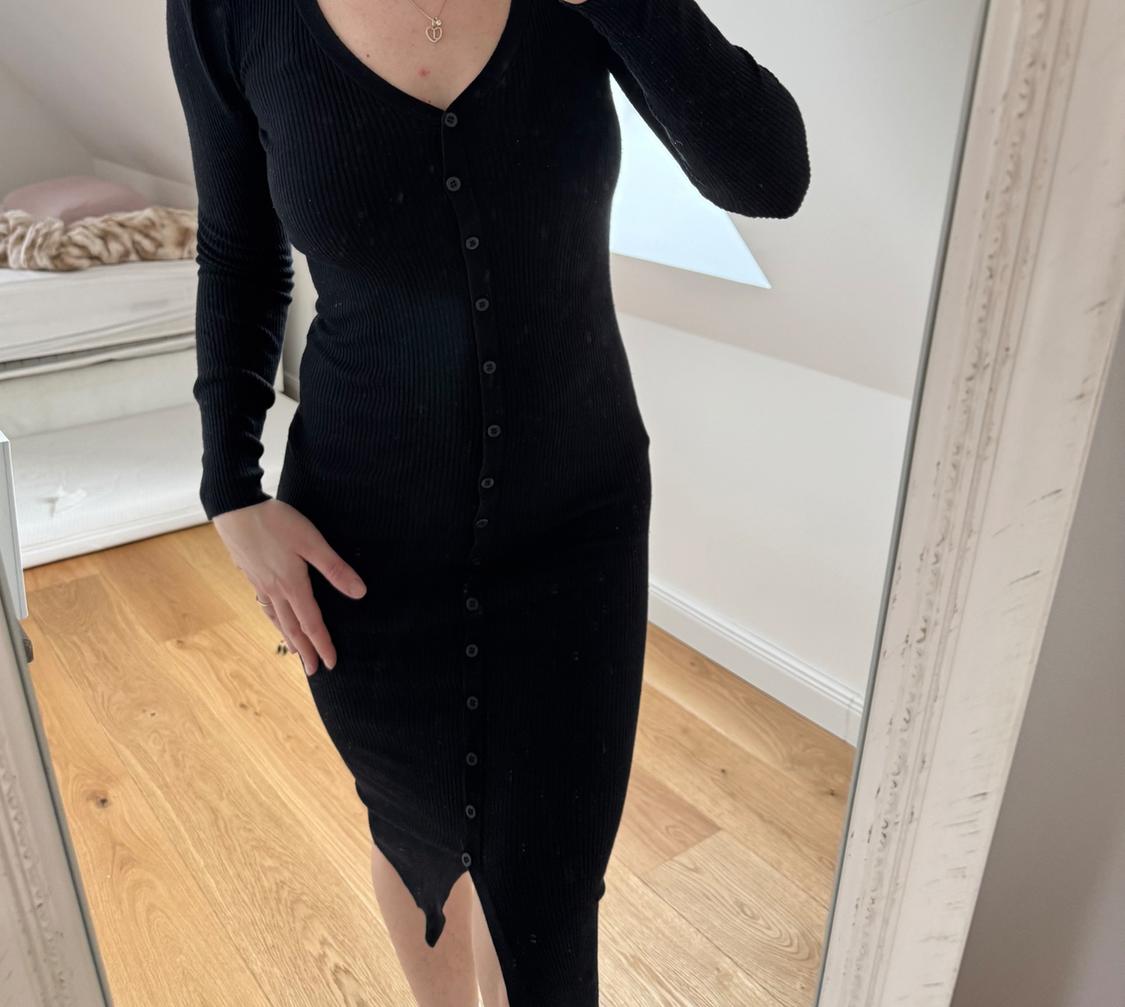 Woman in a fitted black button-up dress, photographed in front of a mirror in a room with a wooden floor and a white bed.